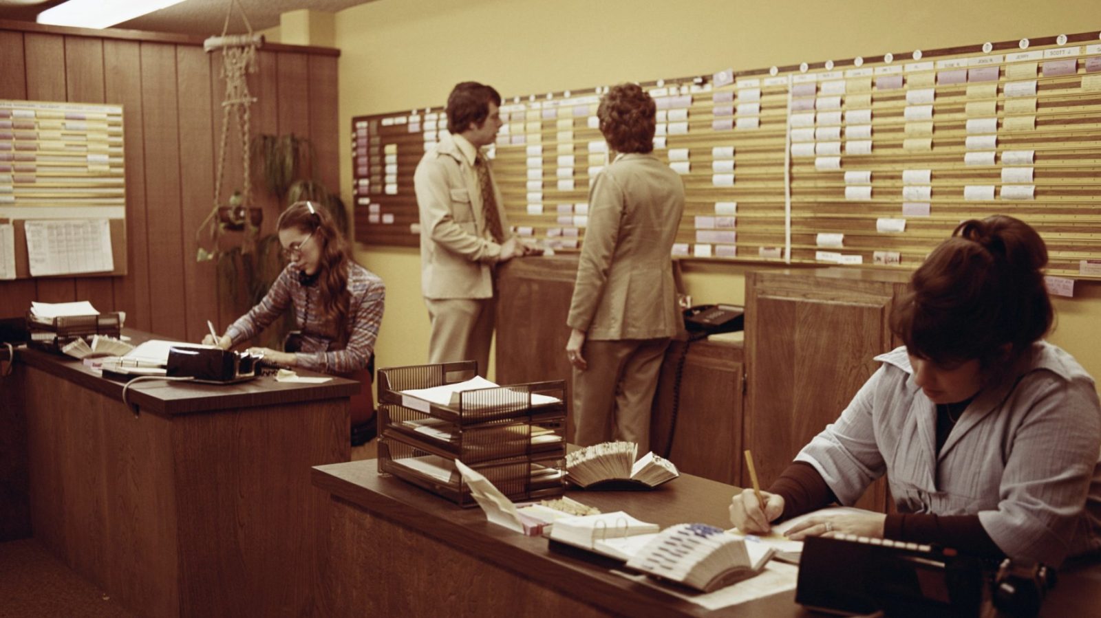 office staff at desks in front of a wall-length scheduling board, 1970s