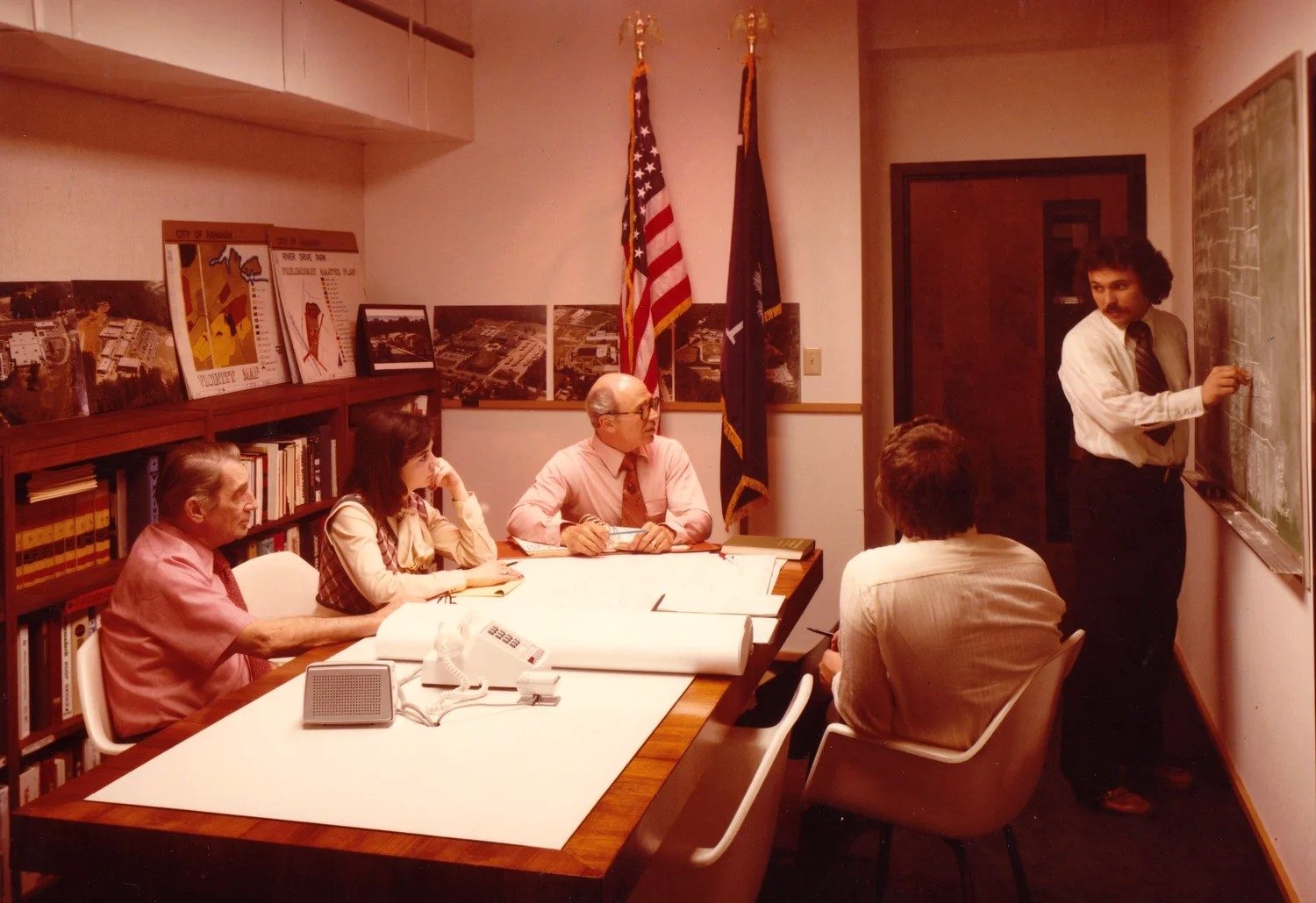 a team meeting around a conference table while a colleague sketches at a chalkboard, 1970s
